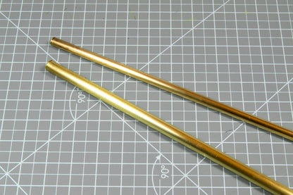 Two brass-colored metal rods, one slightly darker, lie on a gray cutting mat with a grid and 90-degree markings.