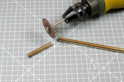 A rotary tool with a cutting disc is positioned near a brass rod, freshly cut into two pieces, on a grid mat