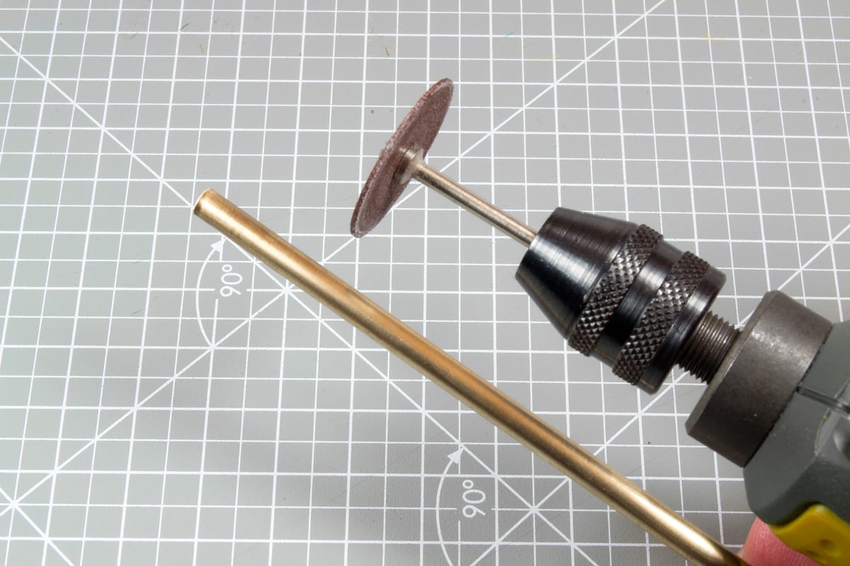 A mini rotary tool with a sanding disc is placed beside a brass rod on a grey grid cutting mat.