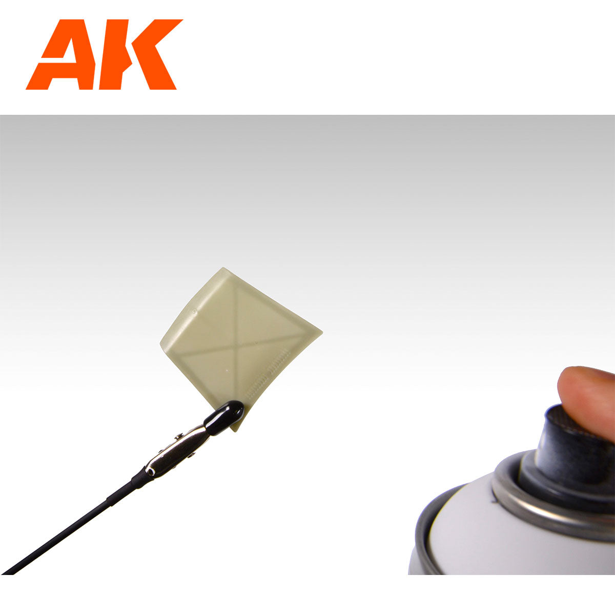 Hand presses spray can nozzle towards a small square model part held by an alligator clip