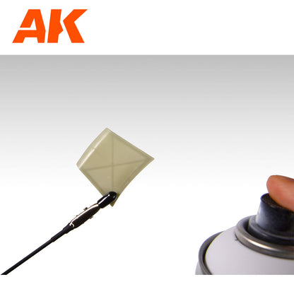 Hand presses spray can nozzle towards a small square model part held by an alligator clip