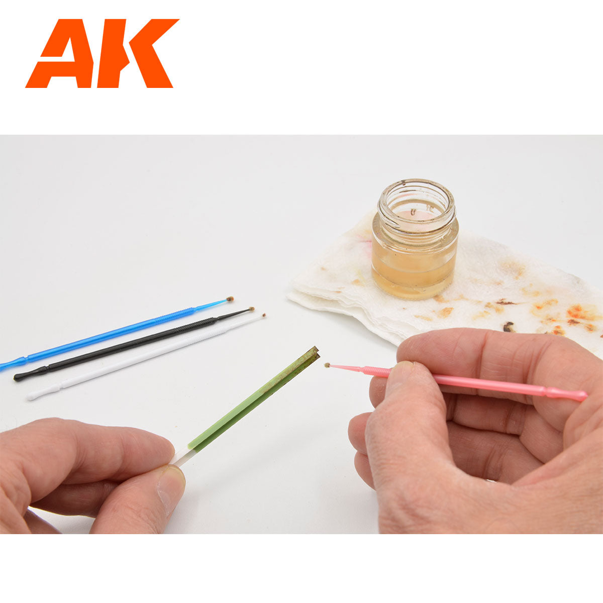 Hands use a pink micro-brush to apply liquid to a green stick. Other brushes and a jar of liquid are on the white surface