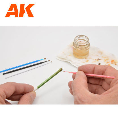 Hands use a pink micro-brush to apply liquid to a green stick. Other brushes and a jar of liquid are on the white surface