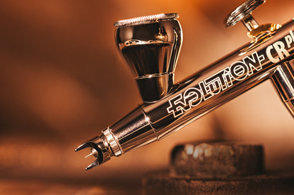 Close-up of a chrome airbrush gun, highlighting the paint cup and fine nozzle tip against a warm background