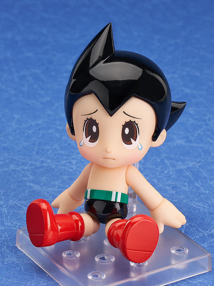 A sad Astro Boy figure sits crying, wearing red boots and black shorts on a clear stand. Blue background