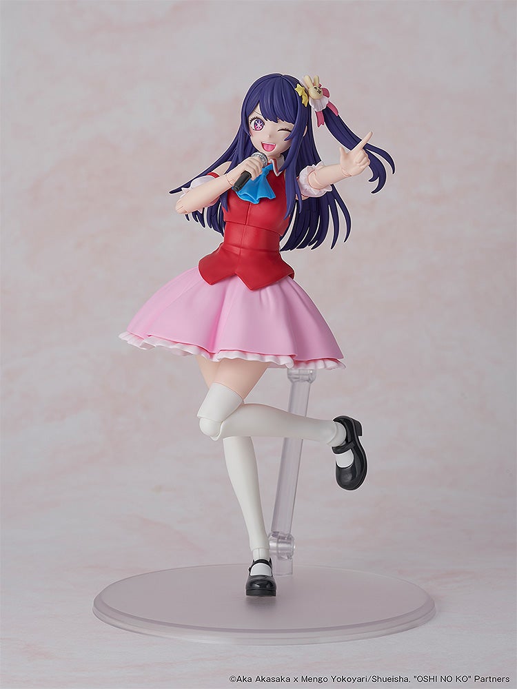 A vibrant anime idol figure with purple hair, winking and holding a mic. She wears a red top, pink skirt, and points excitedly
