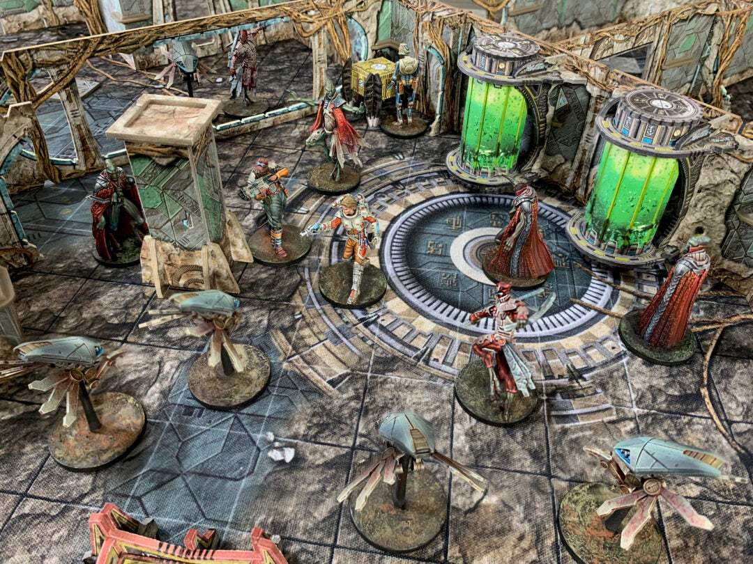 Tabletop sci-fi game with painted miniatures: characters, drones, and robed figures on a ruined map with glowing green tanks