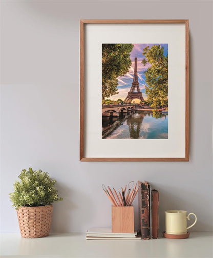 Eiffel Tower rising above a bridge, reflected in water, flanked by trees in a framed picture over a desk.