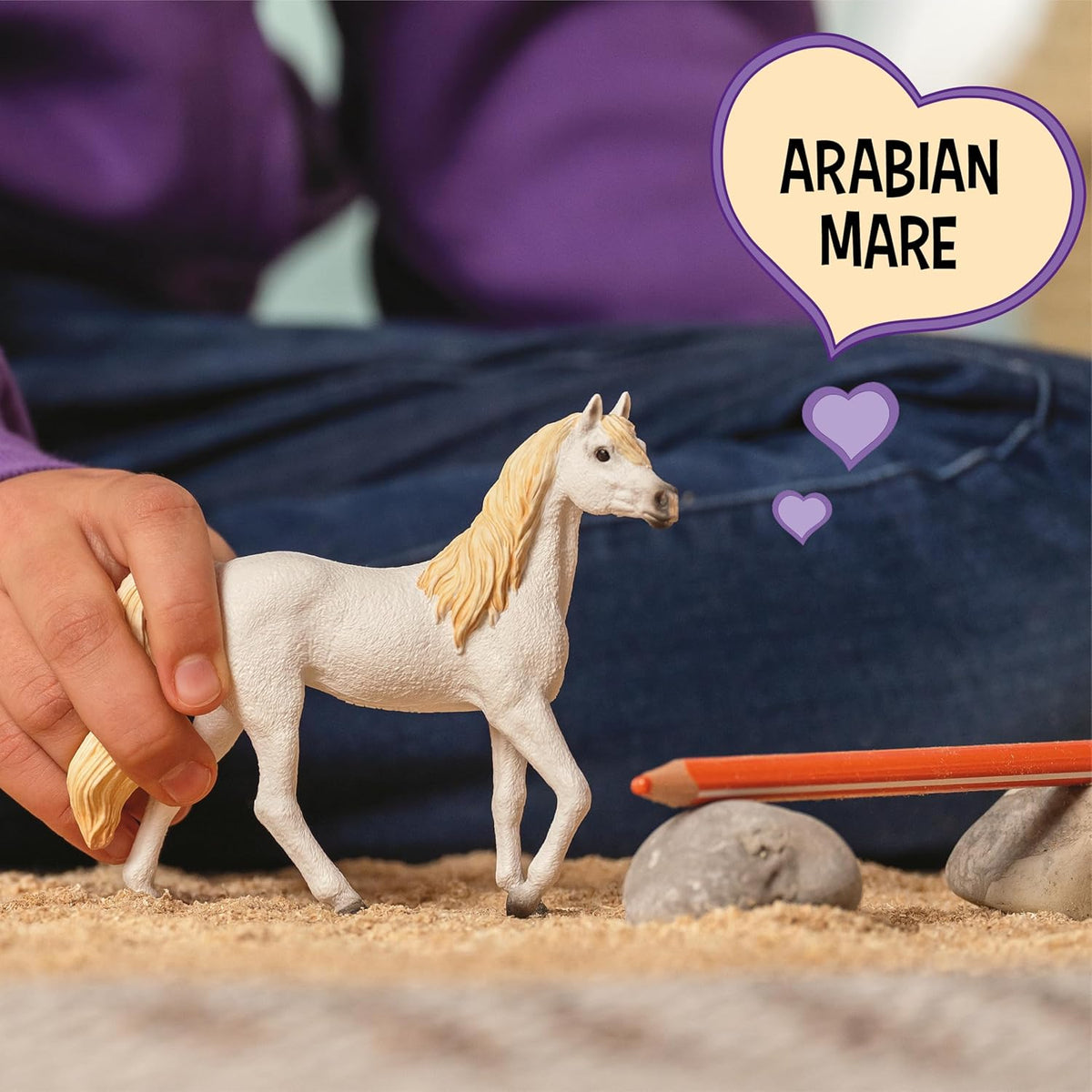 Child plays with a white toy Arabian Mare horse on sand, with a speech bubble reading &#39;ARABIAN MARE&#39; and hearts