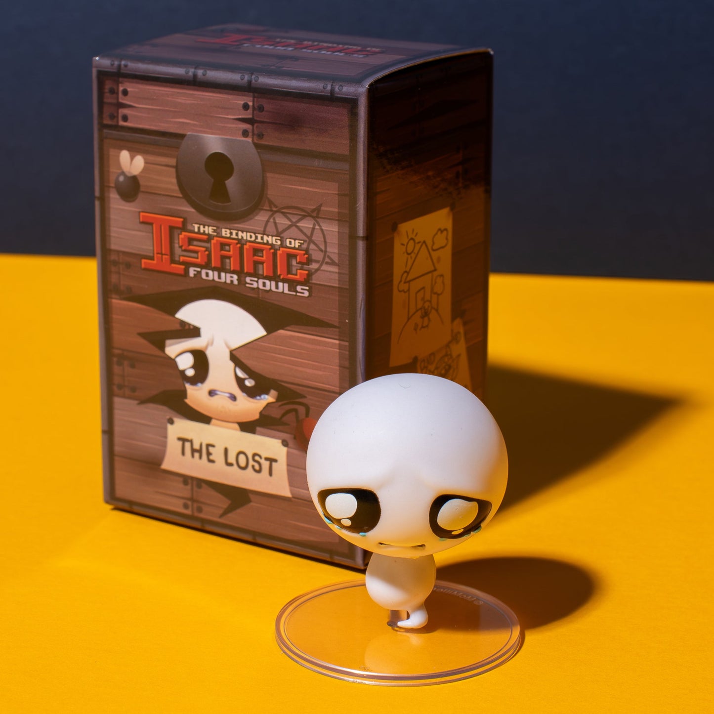 A crying white figure from The Binding of Isaac Collection Figures, named The Lost, stands before its chest-shaped game box
