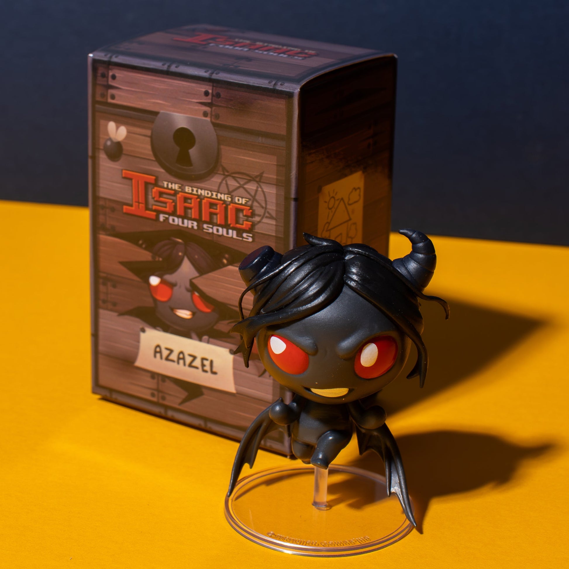 Black Azazel figure with red eyes and horns stands on a clear base in front of its game box for The Binding of Isaac Four Souls