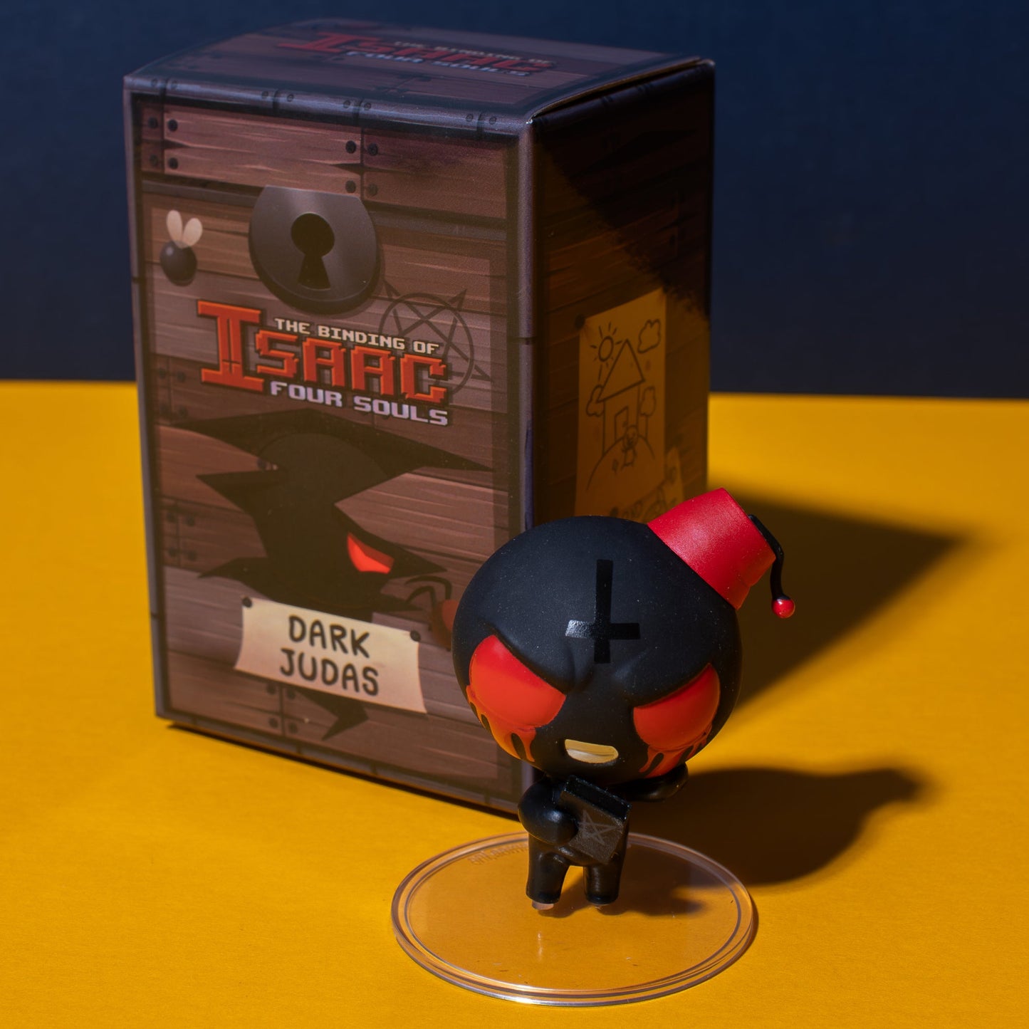 A Dark Judas figure from The Binding of Isaac Collection, with red eyes and a fez, stands on a clear base in front of its chest-like game box