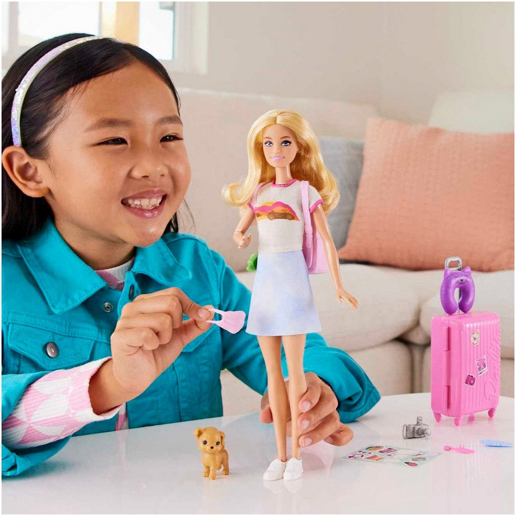 A smiling girl plays with a Barbie doll and travel accessories: pink suitcase, puppy, and camera on a table