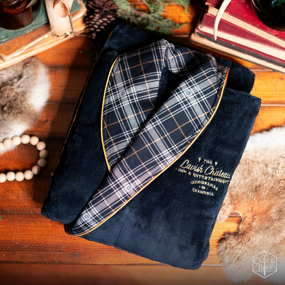 Folded navy blue lavish chateau bathrobe with a plaid collar, gold piping, and embroidered "Lavish Chateau" logo on a wooden surface