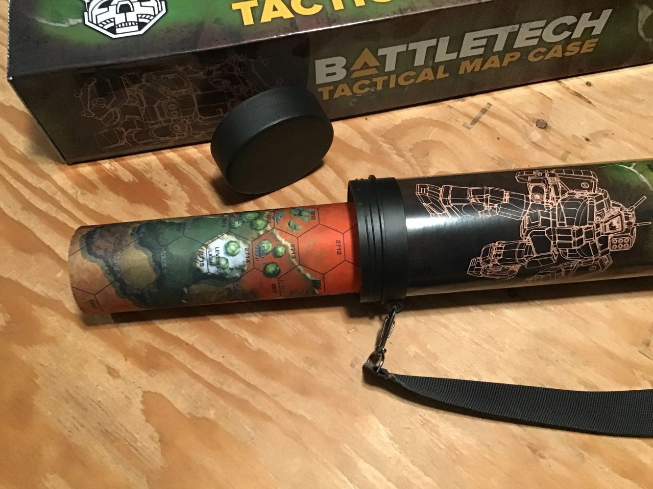 A black BattleTech map case with mech art, partially unrolled to reveal a colorful hex-grid terrain map. Its lid is nearby