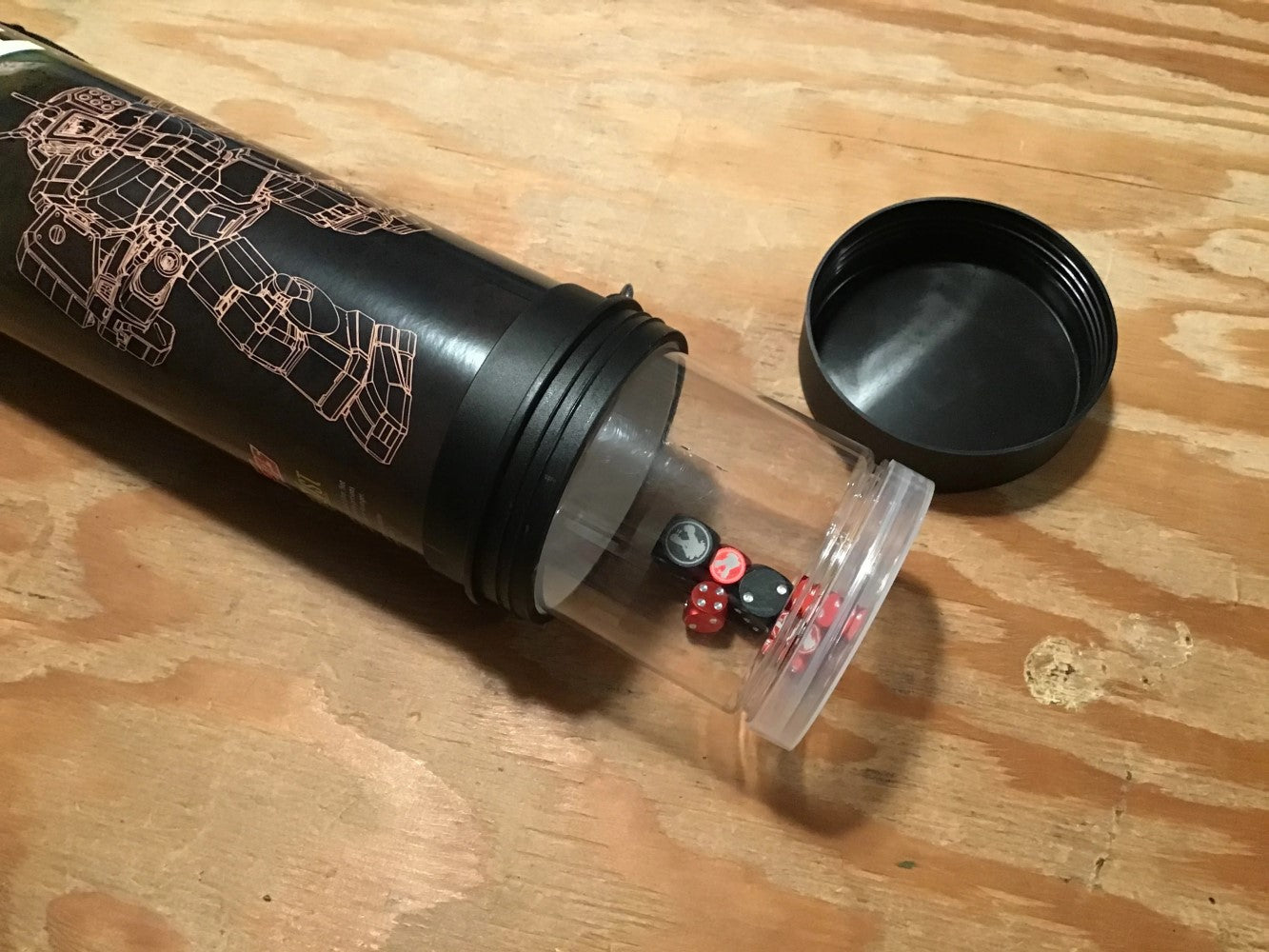 Black cylindrical dice case with a robot graphic, open, revealing red and black dice in its clear end on wood. Lid off