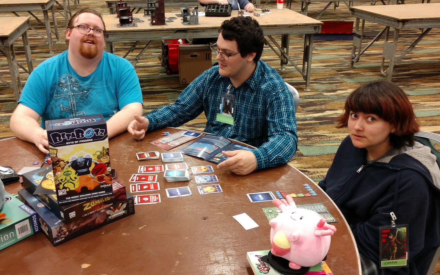 Three people play a card game at a table, surrounded by game boxes, cards, and a pink plush toy.