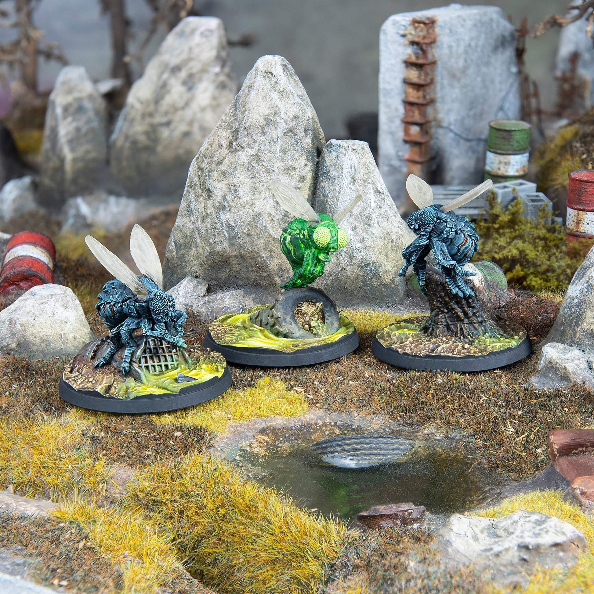 Three mutated fly miniatures stand in a polluted, rocky diorama with rusted debris and vibrant green-yellow toxic waste