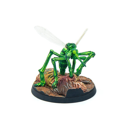 A bright green insect creature miniature, with clear wings, emerges from a brown, organic mound on a black display base