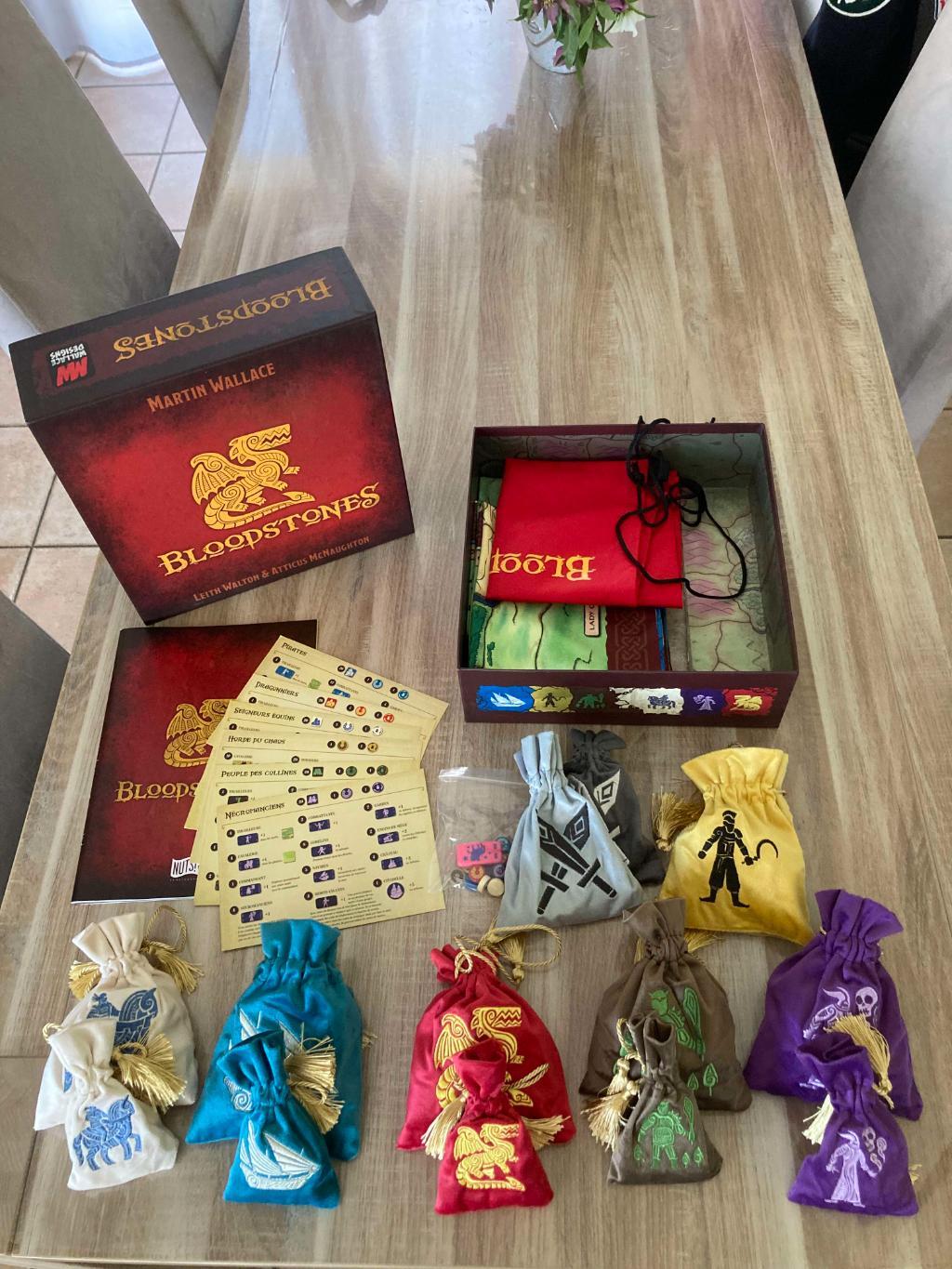 Bloodstones board game components unboxed on a wooden table: red box, rulebooks, and multiple colorful bags with fantasy designs.