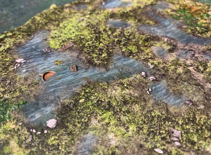 Winding river miniature terrain with green banks, an overturned brown boat, and wooden planks floating in the water.