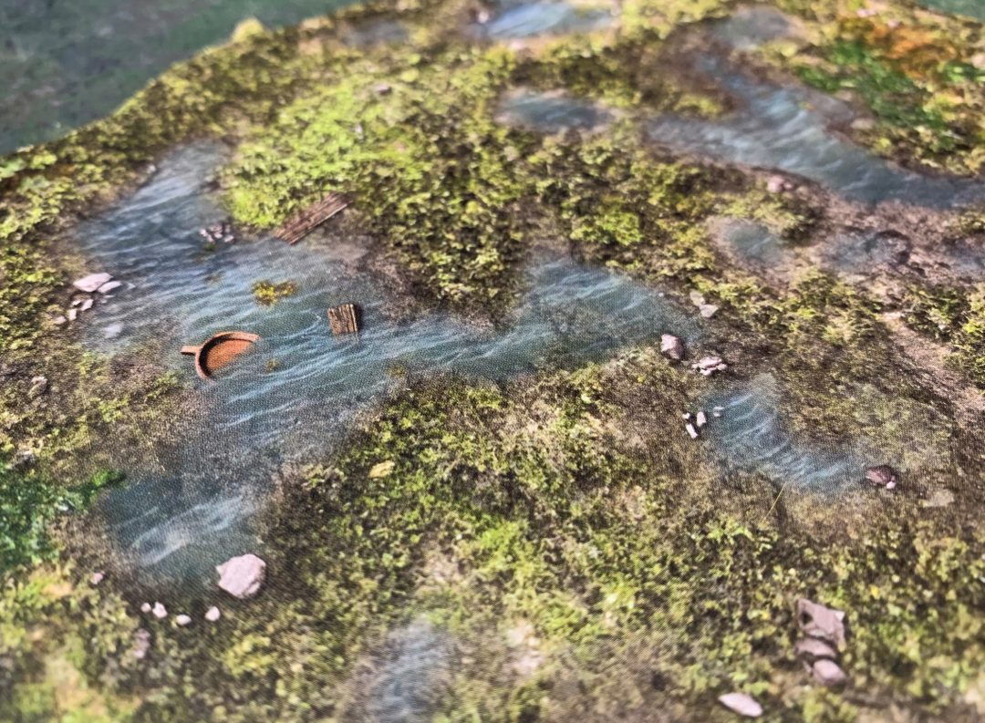 Winding river miniature terrain with green banks, an overturned brown boat, and wooden planks floating in the water.