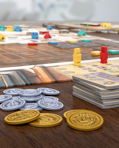 Board game setup with colorful pieces on the board, silver and gold coins, and a stack of cards on a wooden table