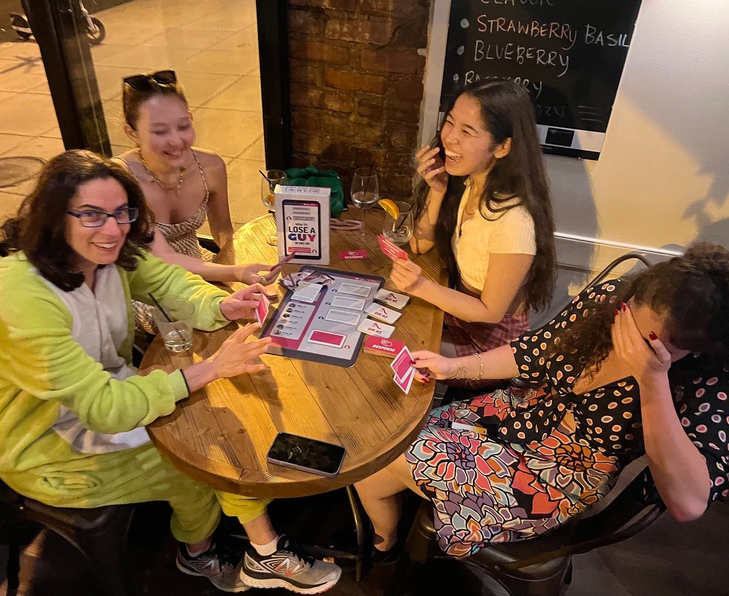 Four friends laugh and interact while playing the How to Lose a Guy in One DM Card Game around a wooden table with drinks