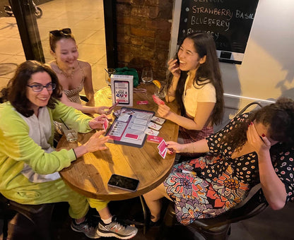 Four friends laugh and interact while playing the How to Lose a Guy in One DM Card Game around a wooden table with drinks