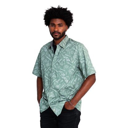 A man with an afro and beard stands, hands in pockets, wearing a sage green shirt patterned with white weapons and symbols