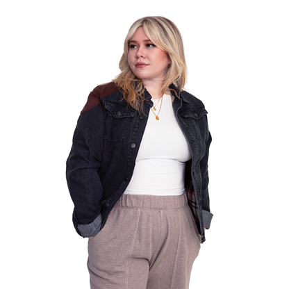 A blonde woman in a dark denim jacket with brown patches and tan sweatpants stands with hands in pockets, looking aside