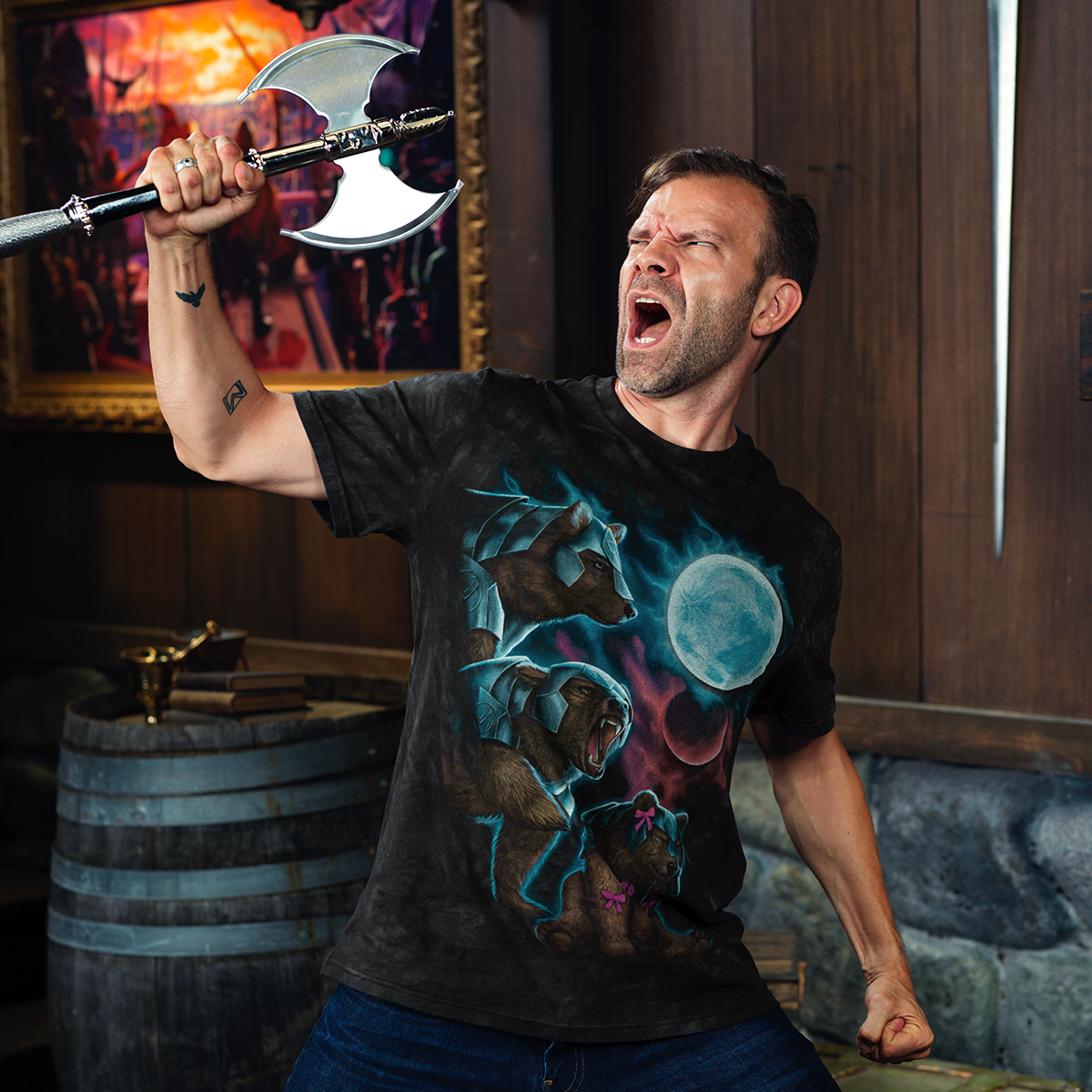A man in an armored bear t-shirt roars, raising a double-bladed axe in a rustic room.