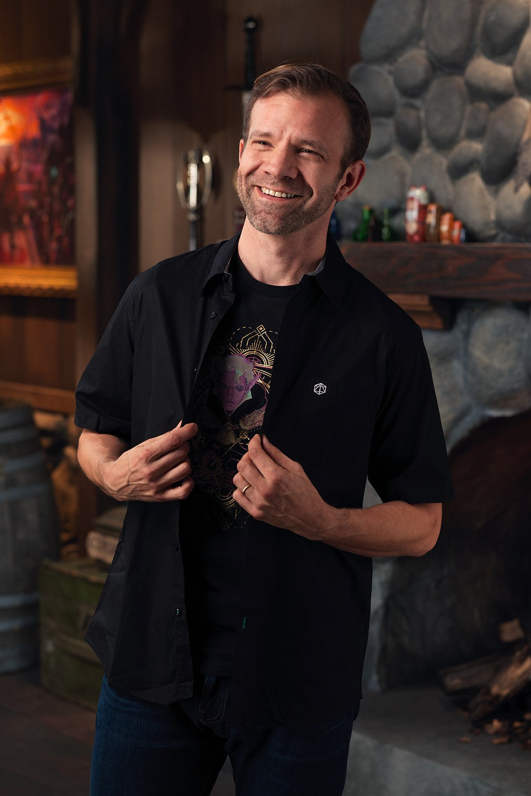 A smiling man holds open his black shirt, revealing a graphic tee with a fantasy character. His outer shirt has a D20 symbol