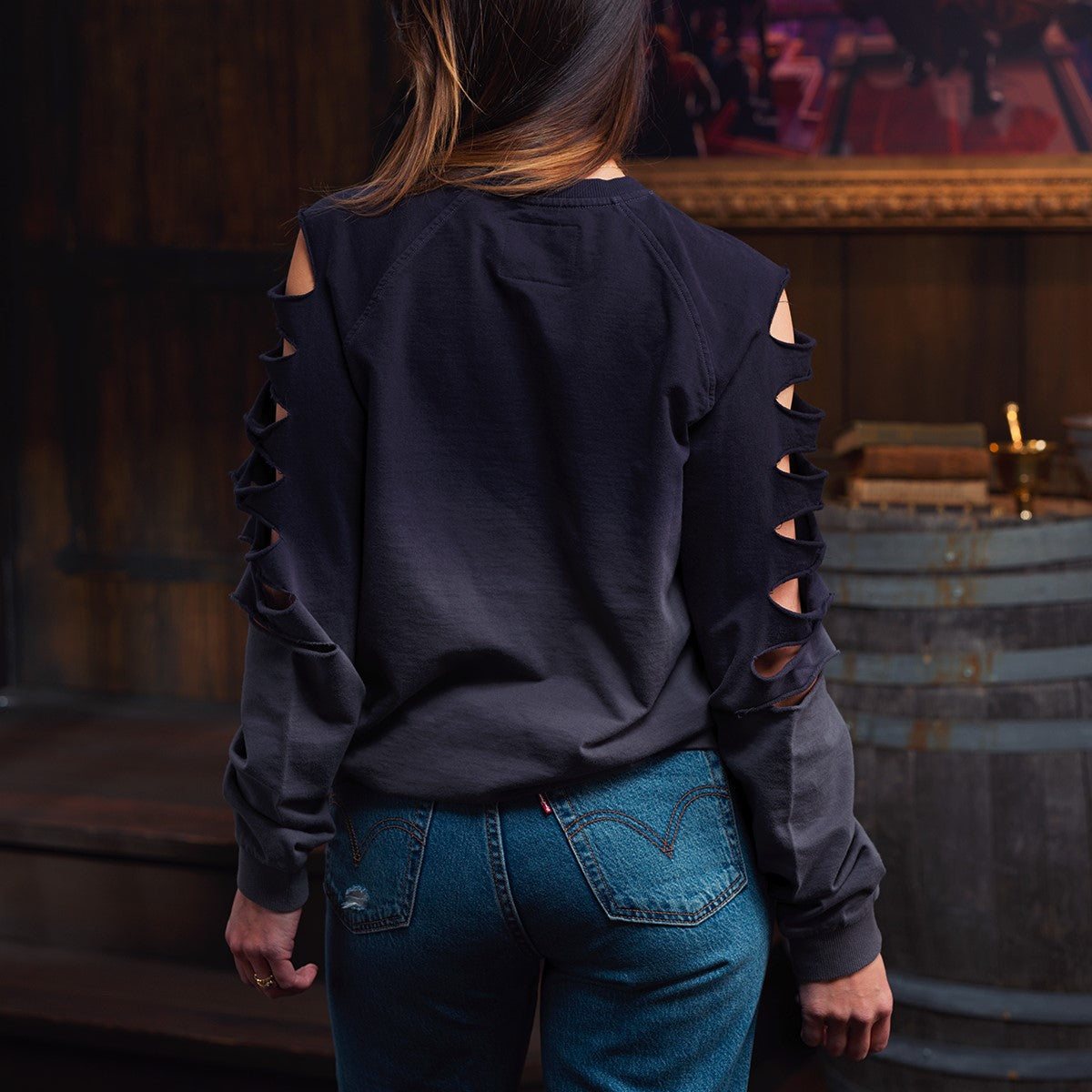 Back view of a girl wearing a dark, long-sleeved crewneck sweatshirt with multiple cutouts along the arms and blue jeans