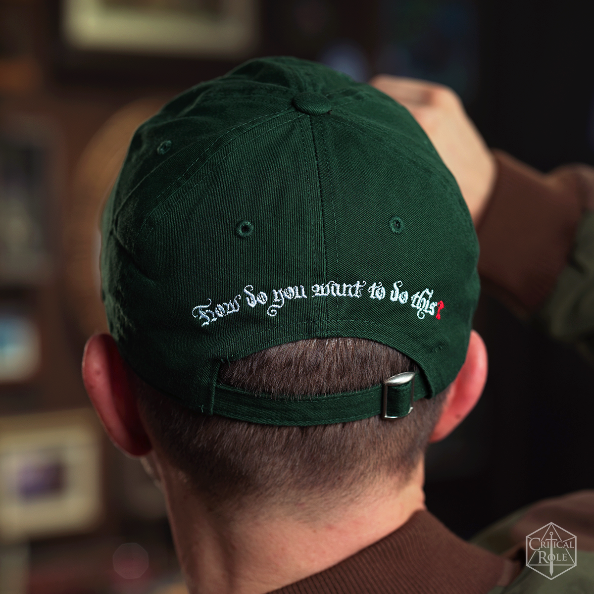 Back of dark green Critical Role Dad hat with white embroidered text: 'How do you want to do this?'. Person's hand adjusts the cap