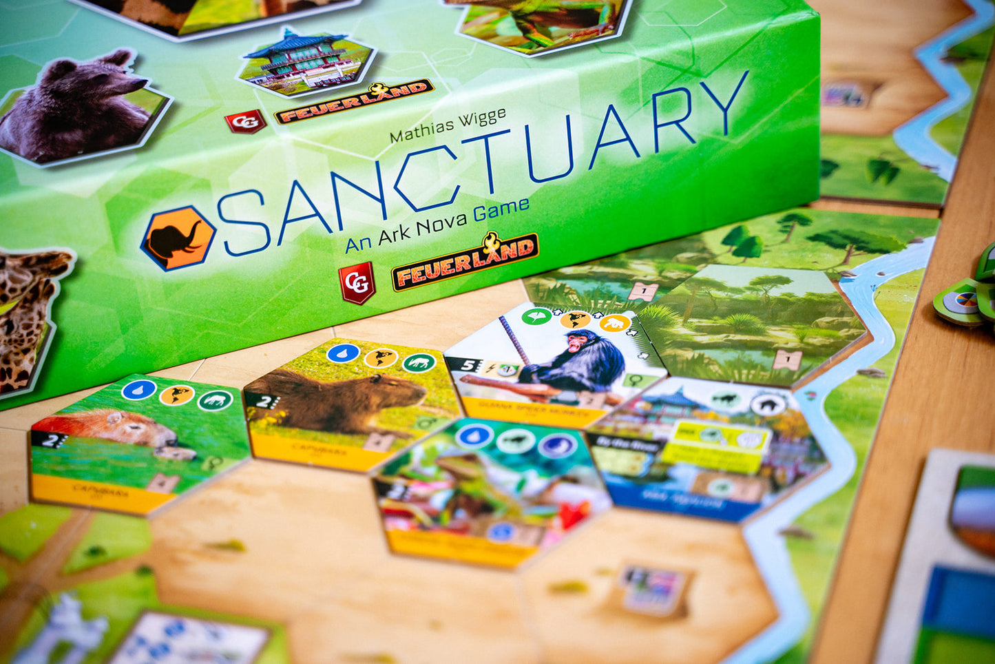 Green box for "Sanctuary: An Ark Nova Game" with hexagonal tiles featuring capybaras, a monkey, and terrain