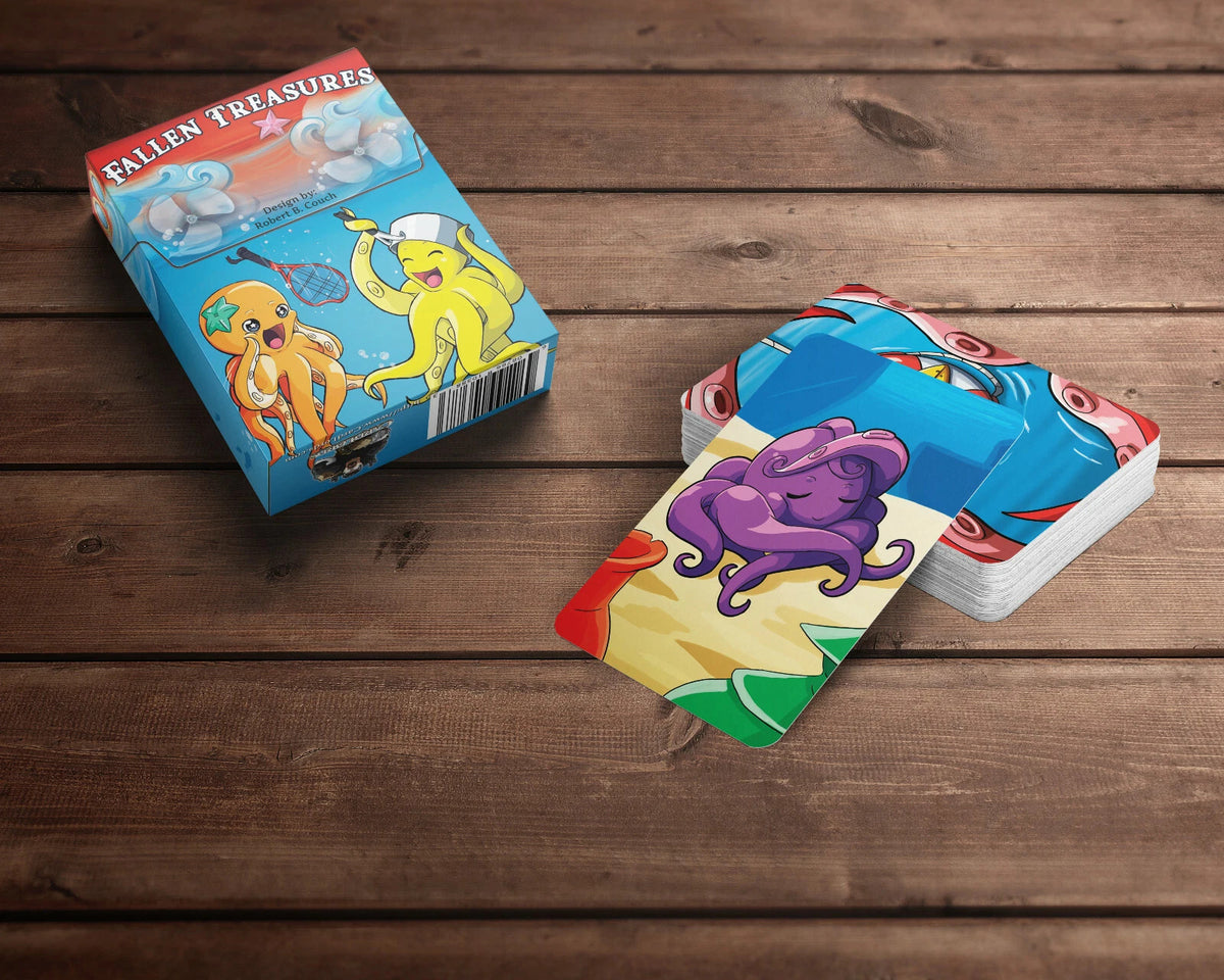 A &quot;Fallen Treasures&quot; card game box featuring two cartoon octopuses, next to a stack of cards with a purple octopus, on wood.