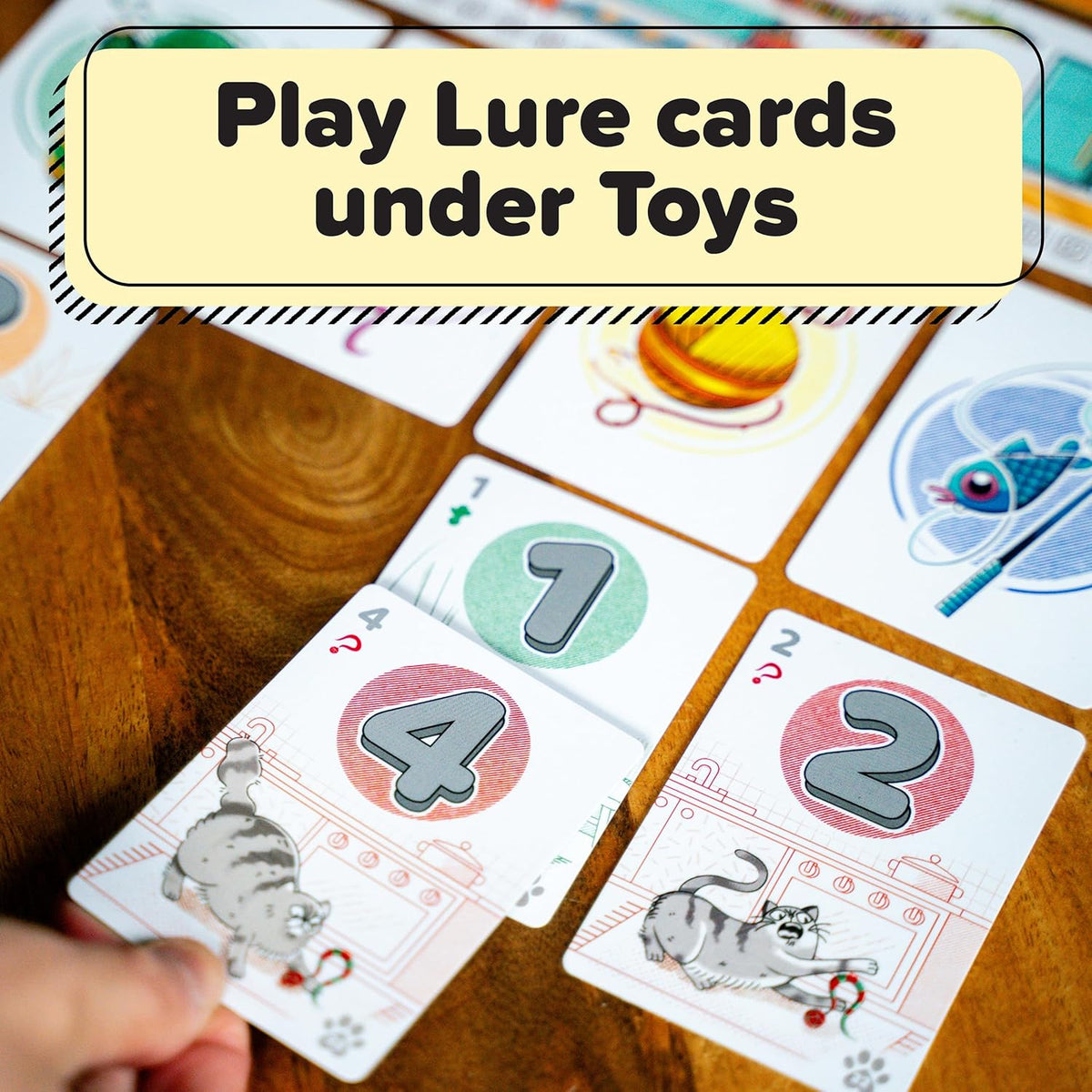 Hand placing cat-themed game cards (numbers 2, 4, 7) on a wooden table. Text: &quot;Play Lure cards under Toys&quot;