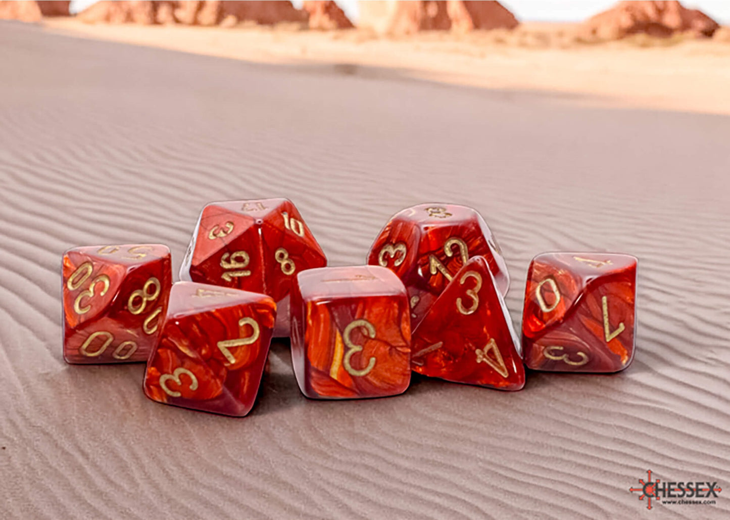 Red marbled polyhedral dice with gold numbers lie scattered on rippled desert sand with distant rocky hills
