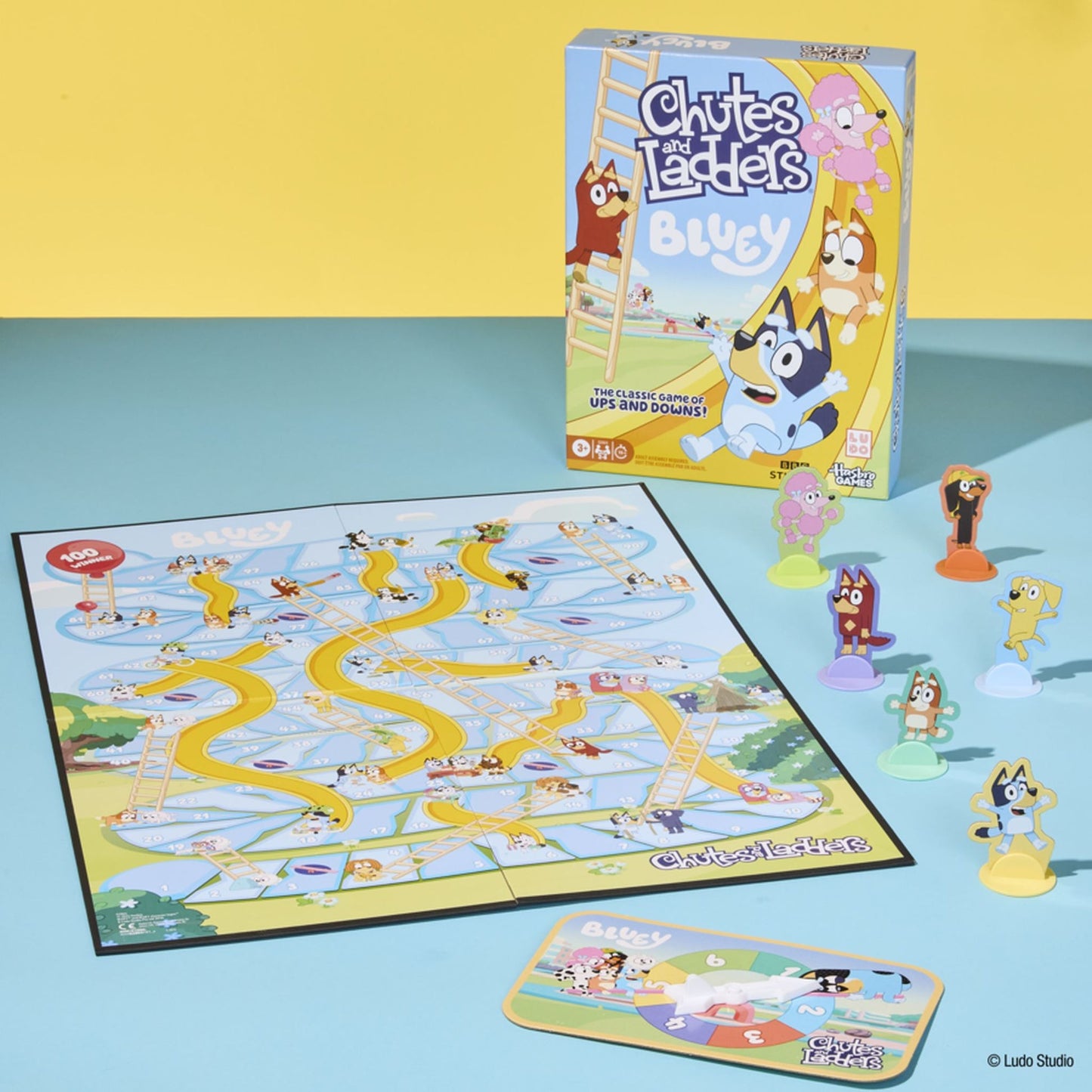 Bluey Chutes and Ladders board game set, with the box, game board, character pawns (including Bluey and Bingo), and a spinner on a light blue table against a yellow wall