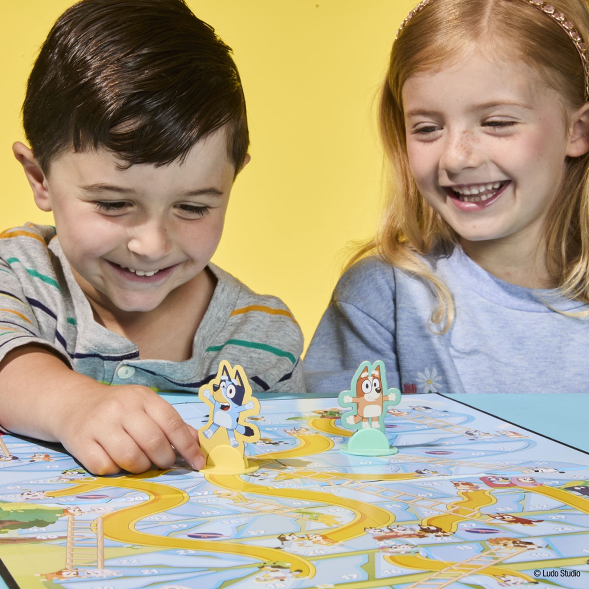 Smiling boy and girl playing a Bluey board game, the boy moves a Bluey character piece along the game board's winding path as the girl laughs happily