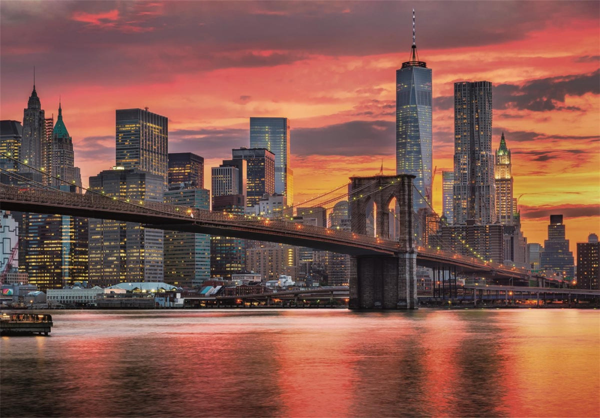 Illuminated Brooklyn Bridge and New York City skyline glow under a vibrant sunset, reflected in the river
