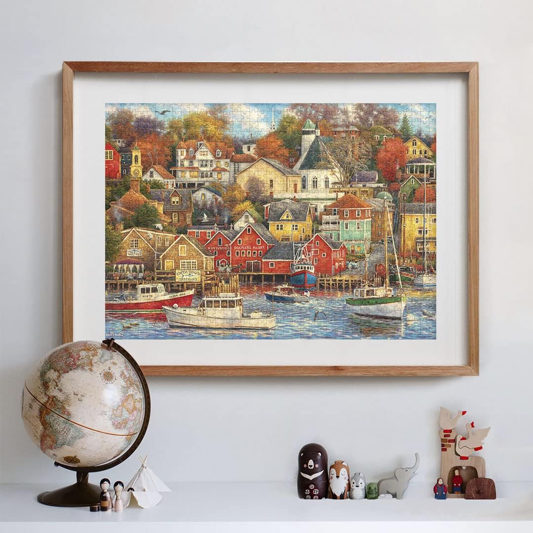 A framed jigsaw puzzle showing a vibrant harbor town with colorful houses, boats, and autumn trees, above a shelf