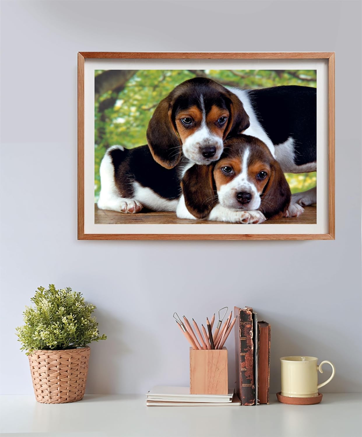A framed photo of two tri-color beagle puppies on a wall above a white desk with a potted plant, books, and pencils