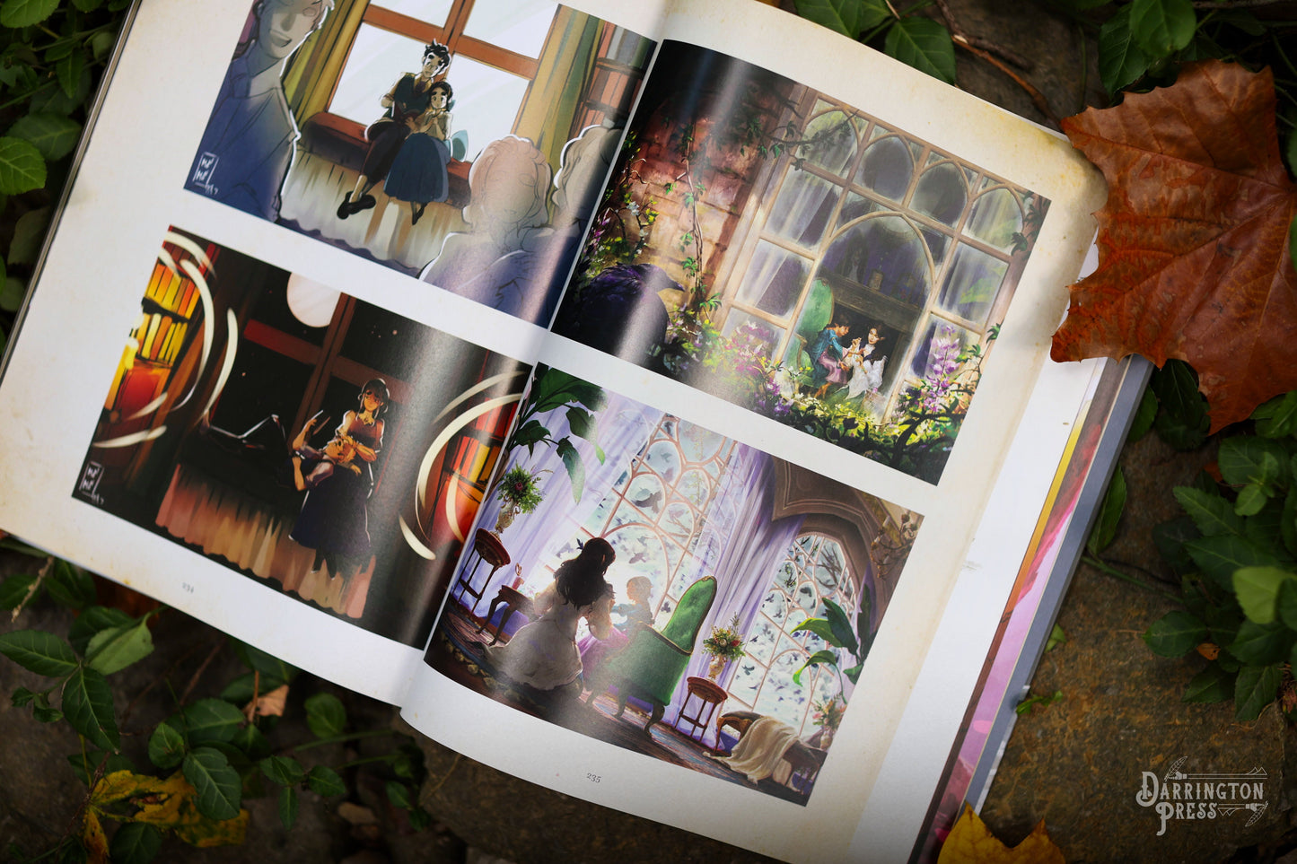 An open art book displays colourful anime-style illustrations of characters in whimsical settings, resting on rocky, leafy ground