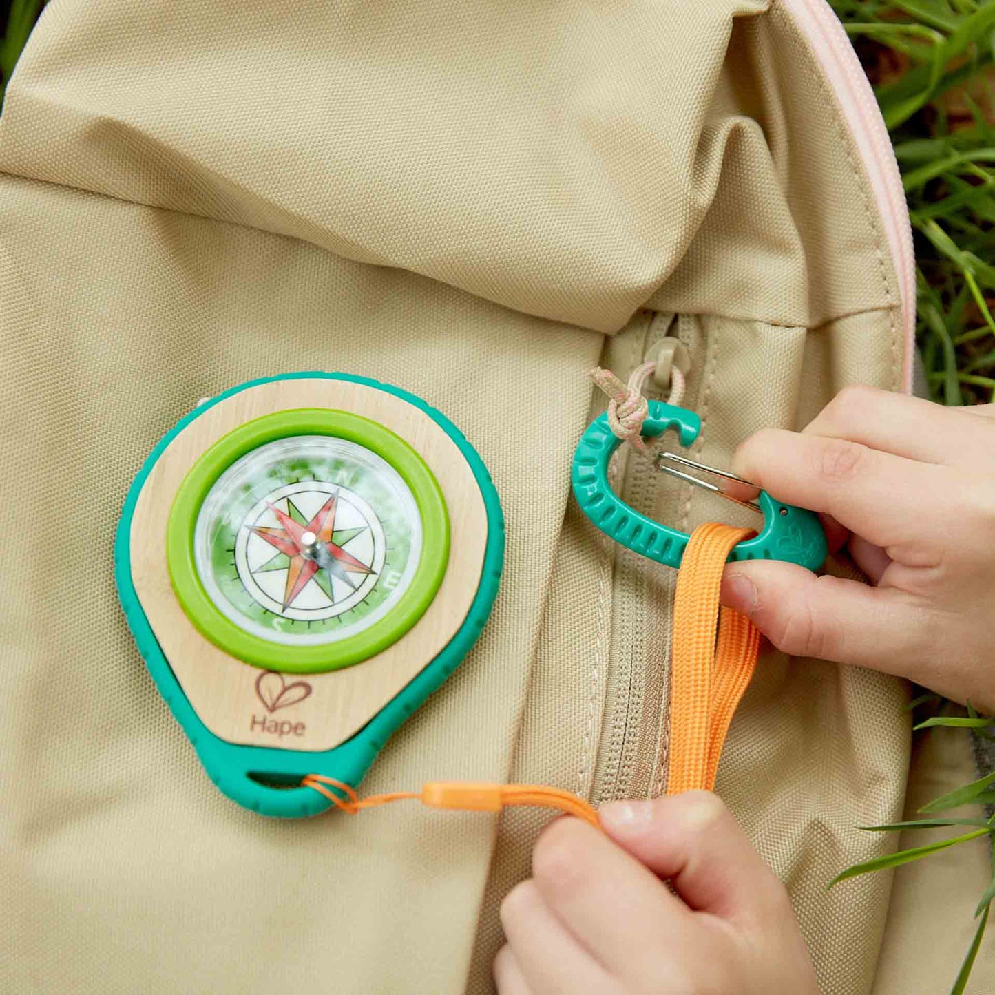Hands clipping a wooden toy compass with an orange lanyard onto a tan backpack