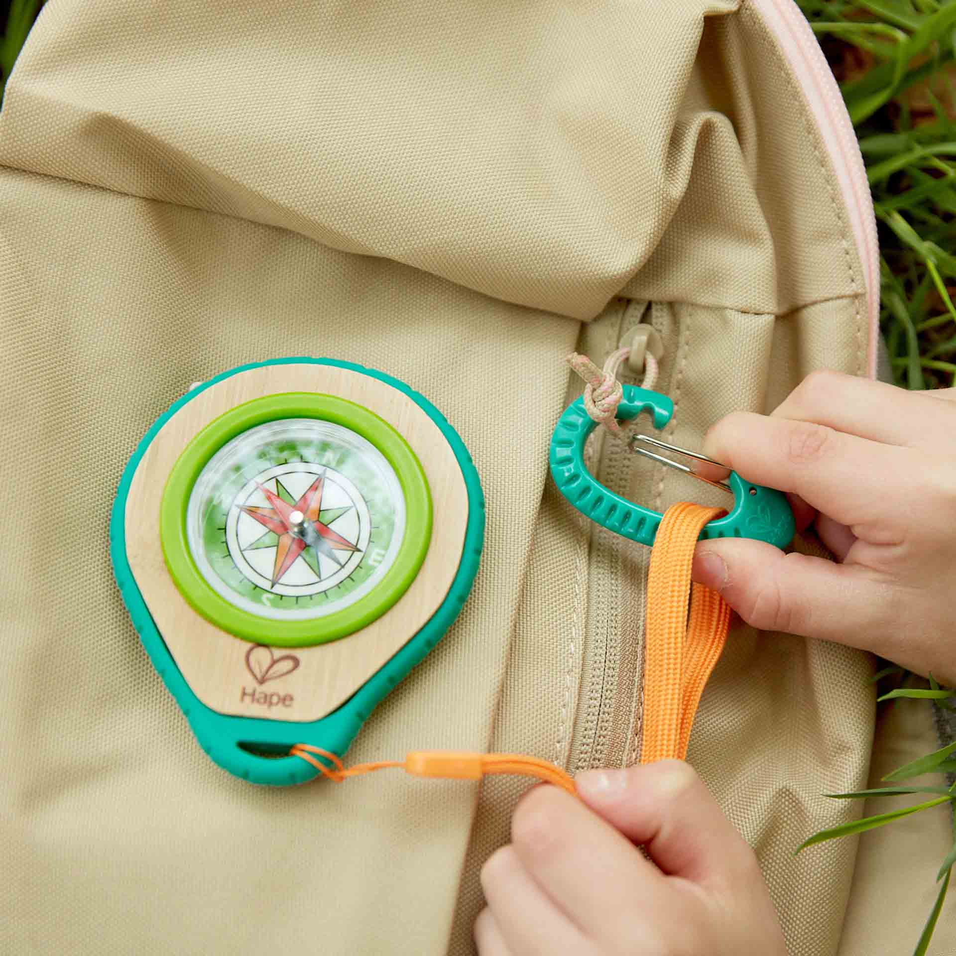 Hands clipping a wooden toy compass with an orange lanyard onto a tan backpack