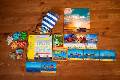The Conservas board game, with its box, rulebook, cards, tokens, and a striped bag, is laid out on a wooden table