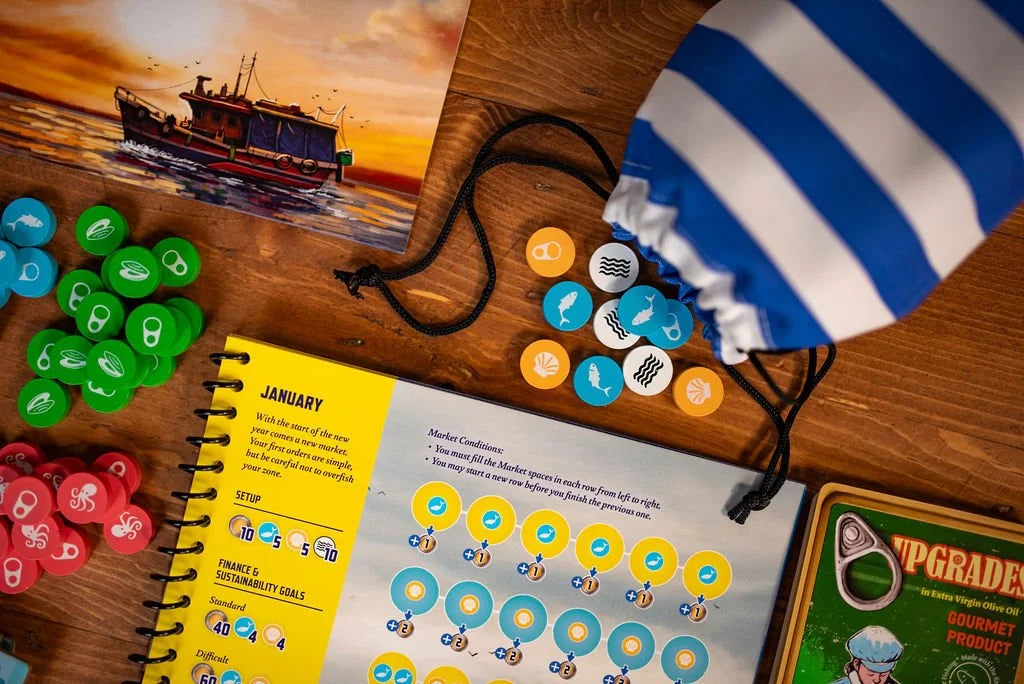 The Conservas board game is set up on a wooden table with colorful tokens, a rulebook open to January, a blue and white striped bag, and a fishing boat illustration