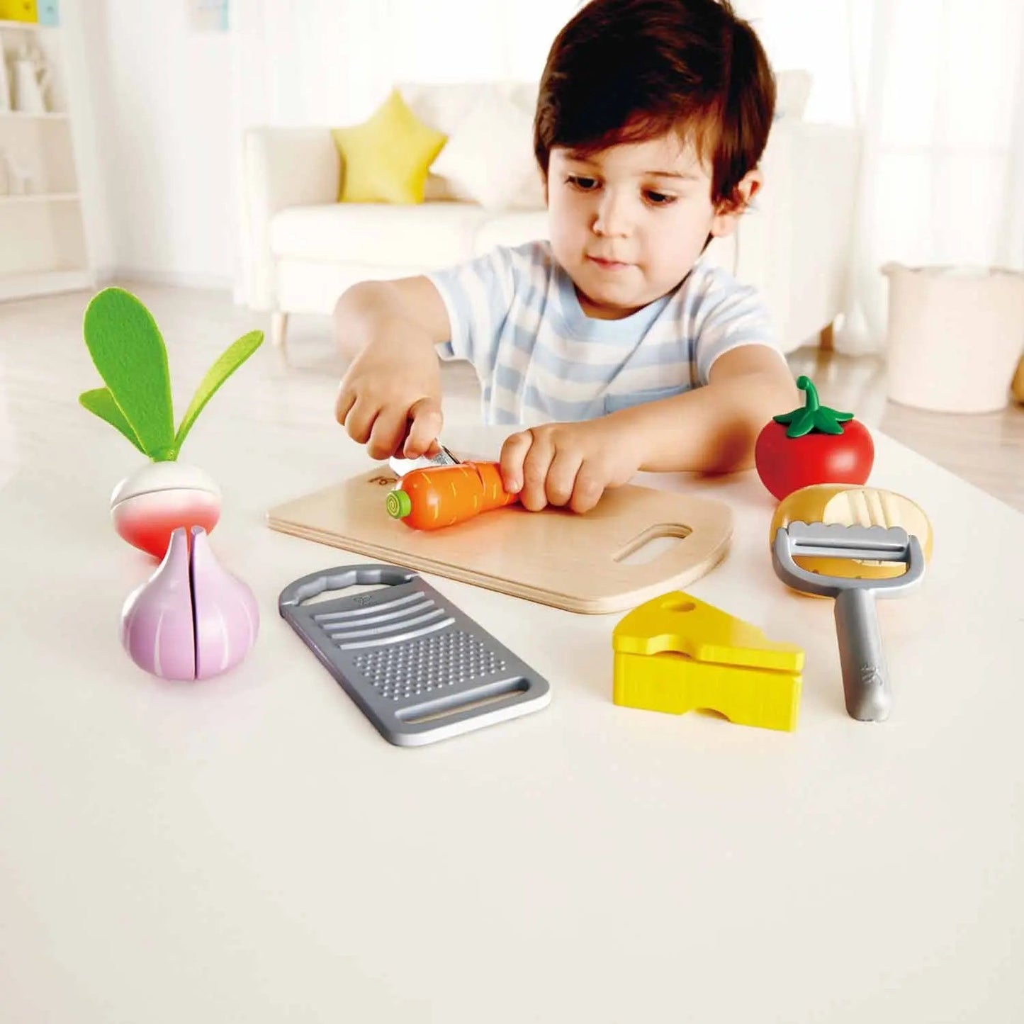 Young boy cutting a toy carrot on a cutting board, surrounded by play food and utensils.